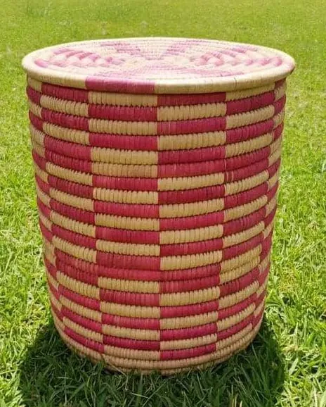 A lidded coiled basket worked in a rose and natural checkerboard pattern, standing on grass